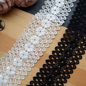 May include: Two lace trims, one white and one black, with a scalloped edge and floral design. The trims are laid out on a wooden surface.