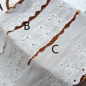 Peut inclure: Trois galons de dentelle en coton blanc avec des bords festonn&eacute;s et des motifs d'ours en peluche brod&eacute;s. Les galons de dentelle sont dispos&eacute;s en pile, le galon du haut pr&eacute;sentant un motif d'ours en peluche plus petit, le galon du milieu pr&eacute;sentant un motif d'ours en peluche plus grand et le galon du bas pr&eacute;sentant un motif d'ours en peluche avec un n&oelig;ud.