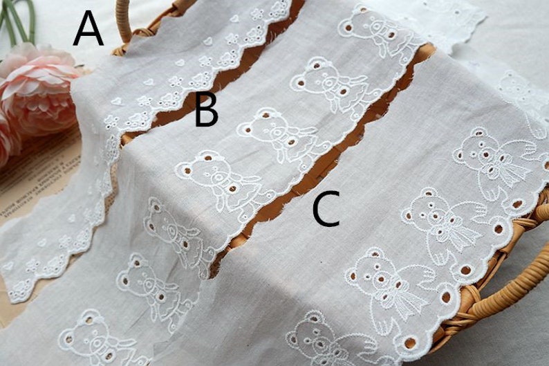 Peut inclure: Trois galons de dentelle en coton blanc avec des bords festonn&eacute;s et des motifs d'ours en peluche brod&eacute;s. Les galons de dentelle sont dispos&eacute;s en pile, le galon du haut pr&eacute;sentant un motif d'ours en peluche plus petit, le galon du milieu pr&eacute;sentant un motif d'ours en peluche plus grand et le galon du bas pr&eacute;sentant un motif d'ours en peluche avec un n&oelig;ud.