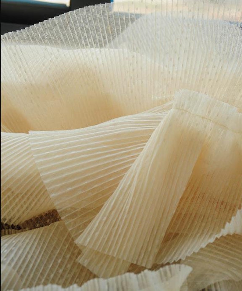 May include: Close-up of a beige pleated fabric with a subtle dotted pattern. The fabric is soft and delicate, with a flowing, layered texture.