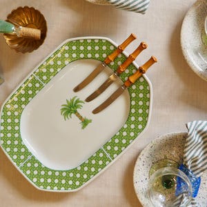 May include: A white oval serving platter with a green and white polka dot pattern. The platter features a palm tree design and four wooden handled knives.