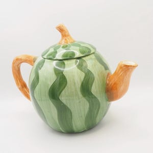 Può includere: Una teiera in ceramica con un design a forma di anguria. Il corpo rotondo è dipinto di verde con strisce ondulate verde scuro. Il manico, il beccuccio e lo stelo del coperchio sono arancioni. Il coperchio è verde con macchie verdi più scure.