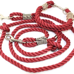 Op de afbeelding: Rode hondenriem en halsband set. De riem en halsband zijn gemaakt van dik, gedraaid touw. De halsband heeft een metalen sluiting en decoratieve secties met zwart-witte accenten. De riem heeft een metalen sluiting en een metalen ring.