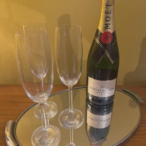Könnte beinhalten: Eine Flasche Moët & Chandon Brut Champagner und drei Champagnerflöten auf einem ovalen Spiegel Tablett. Die Champagnerflasche ist grün mit Goldfolie und einem roten Siegel. Die Flöten sind aus klarem Glas.