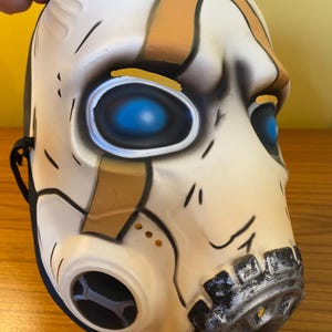 May include: A white and brown plastic mask with blue eyes and a black gas mask. The mask has a distressed look with black and silver accents.