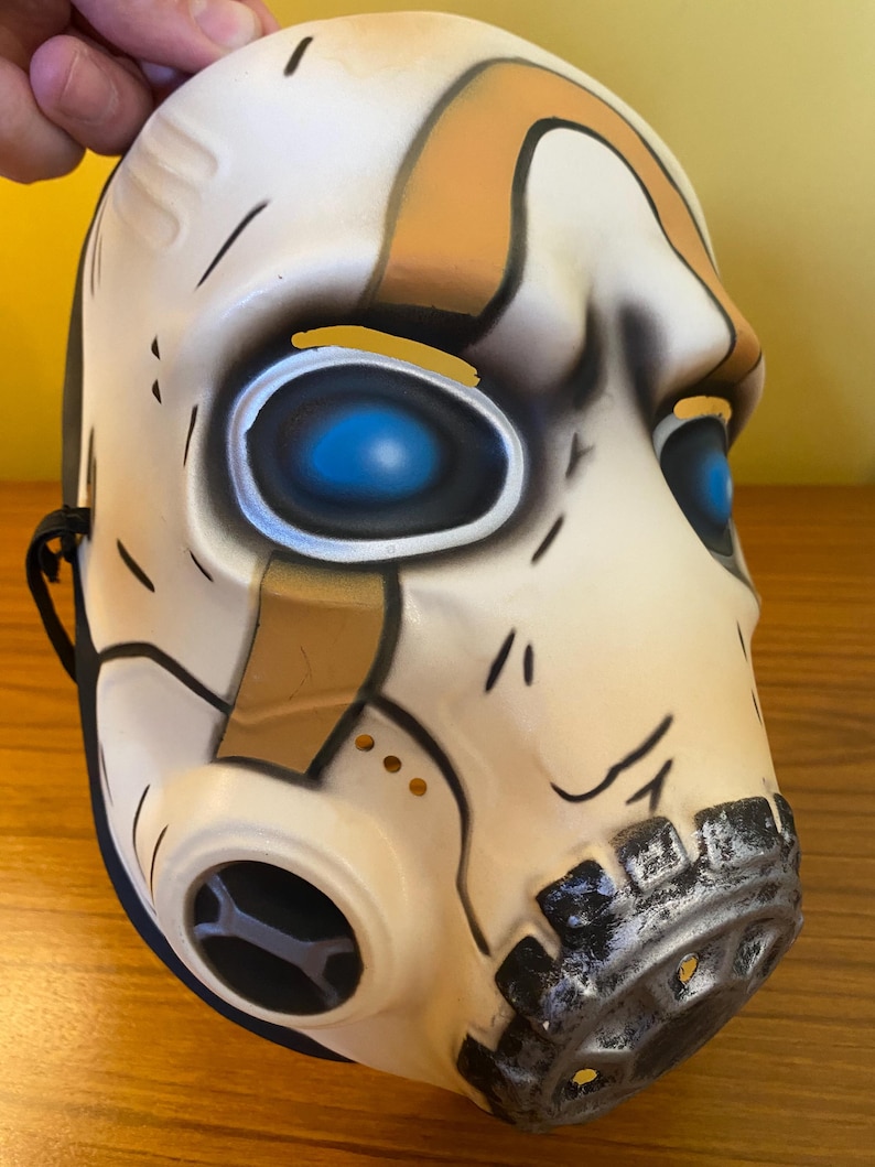 May include: A white and brown plastic mask with blue eyes and a black gas mask. The mask has a distressed look with black and silver accents.
