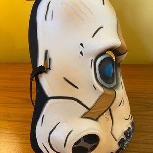 May include: A white plastic skull mask with a black elastic band. The mask has a blue eye and a brown and black gas mask design.