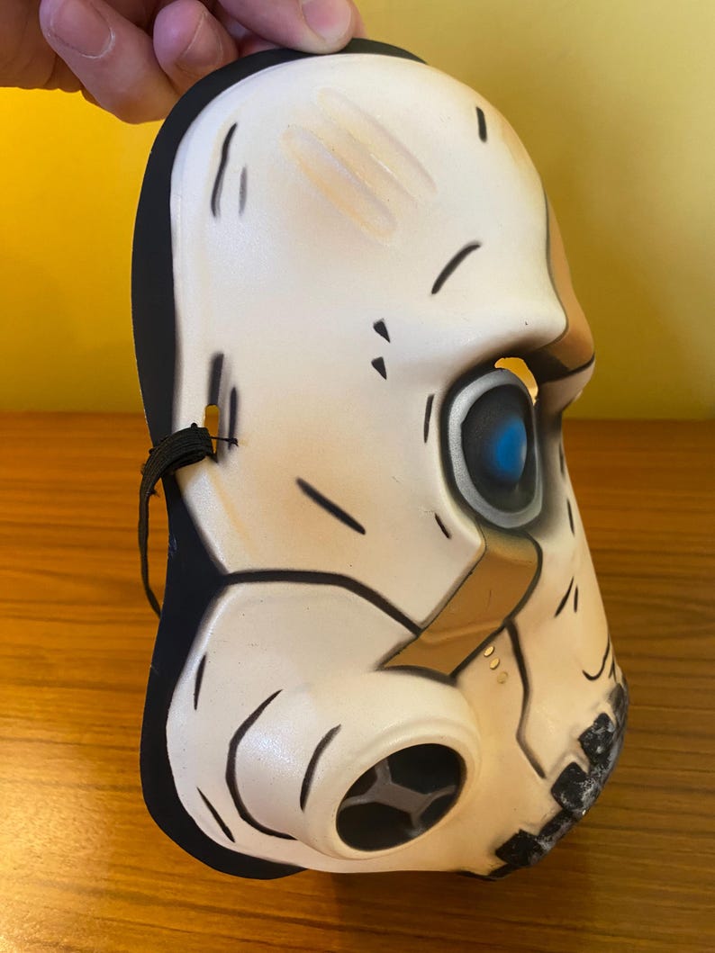 May include: A white plastic skull mask with a black elastic band. The mask has a blue eye and a brown and black gas mask design.
