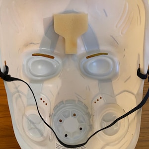 May include: White plastic mask with a black elastic strap. The mask has two eye holes, a nose hole, and a mouth hole. The mask is decorated with a gray and white pattern. The text on the mask reads: "Recommended to reduce wear. Avoid visibility created by unrestricted use. Do not wear near heat or open flame. 2019 Phenom, LLC."