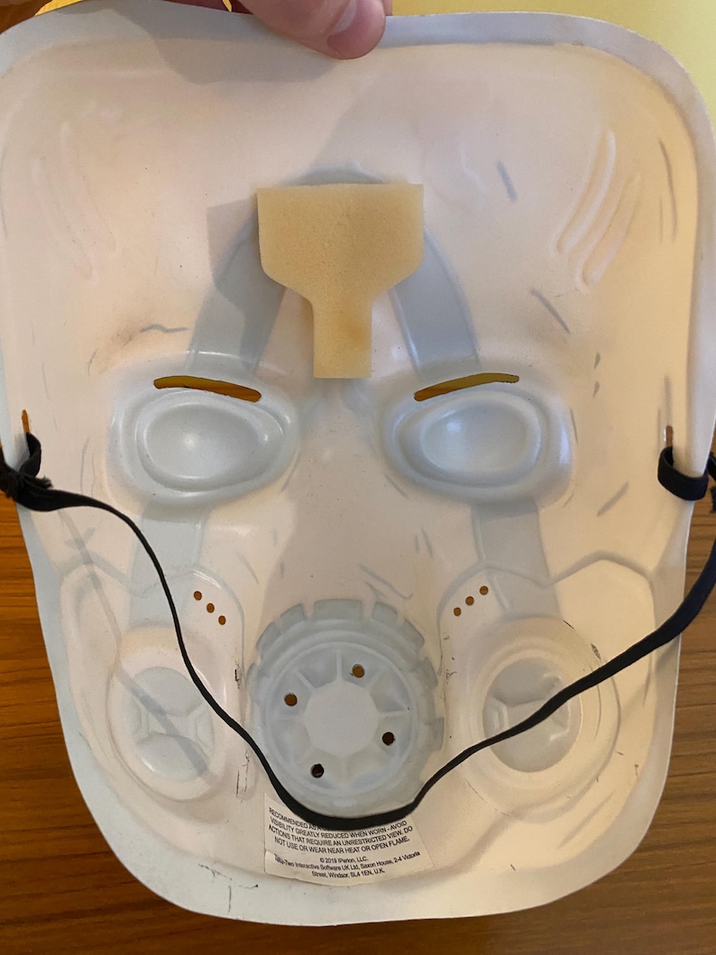 May include: White plastic mask with a black elastic strap. The mask has two eye holes, a nose hole, and a mouth hole. The mask is decorated with a gray and white pattern. The text on the mask reads: "Recommended to reduce wear. Avoid visibility created by unrestricted use. Do not wear near heat or open flame. 2019 Phenom, LLC."