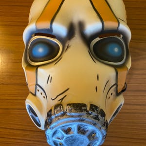 May include: A white plastic mask with orange and black stripes, blue eyes, and a black and gray gas mask mouthpiece.