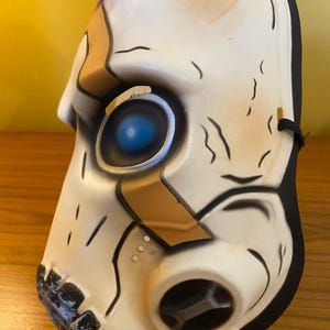 May include: A white plastic mask with a black elastic band. The mask has a gold and black design with a blue eye. The mask is a popular character from the video game Borderlands.