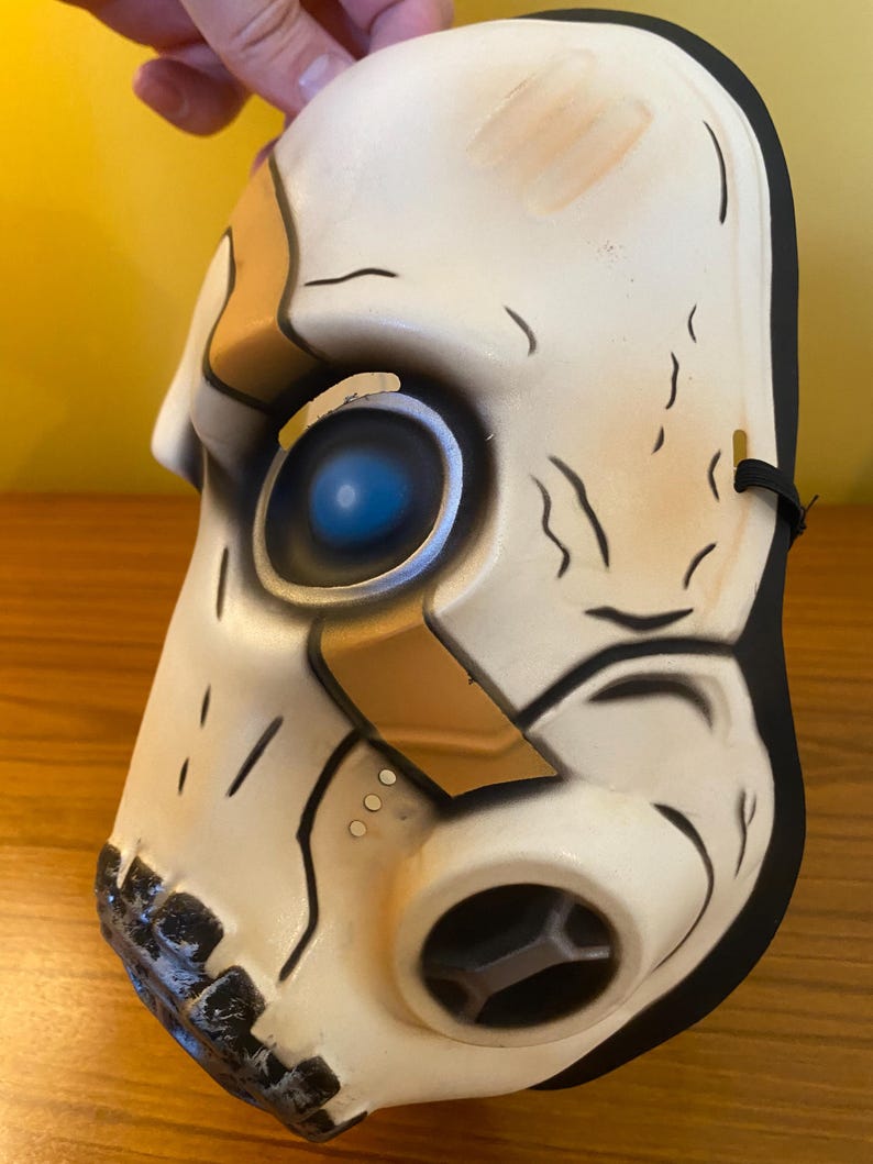 May include: A white plastic mask with a black elastic band. The mask has a gold and black design with a blue eye. The mask is a popular character from the video game Borderlands.