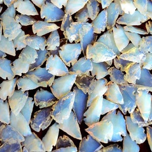 May include: A collection of numerous translucent, pale blue arrowhead-shaped stones. Each piece features a smooth, polished surface with an iridescent sheen that reflects light, creating a layered, mosaic-like arrangement of light blue shapes.