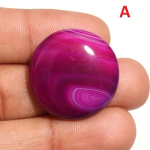Puede incluir: Un cabujón redondo de ágata rosa con bandas blancas. La piedra tiene una superficie lisa y pulida y mide aproximadamente 25 mm de diámetro.