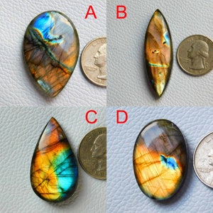 Puede incluir: Cuatro cabujones de labradorita en diferentes formas y tamaños. Las piedras son iridiscentes con tonos de azul, verde, dorado y marrón. Las piedras se muestran junto a monedas de 25 centavos de dólar estadounidense para comparar el tamaño.