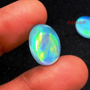 May include: A close-up of a blue, green, and yellow iridescent oval opal gemstone. The opal is being held by a finger.