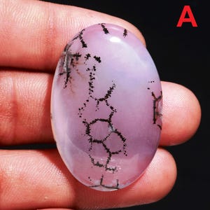May include: An oval gemstone, polished to a high sheen, displaying a soft pink colour with striking black dendritic patterns. The stone is held in fingers, highlighting its smooth texture and unique natural design. The letter "A" is visible.