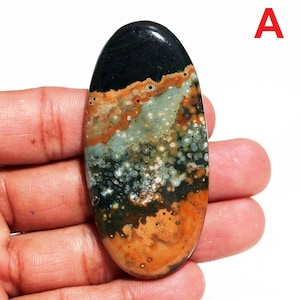 Peut inclure: Une pierre précieuse ovale polie. Le haut est noir, se fondant dans des motifs mouchetés orange, vert et blanc. La pierre est tenue dans une main, mettant en valeur ses couleurs et textures naturelles.
