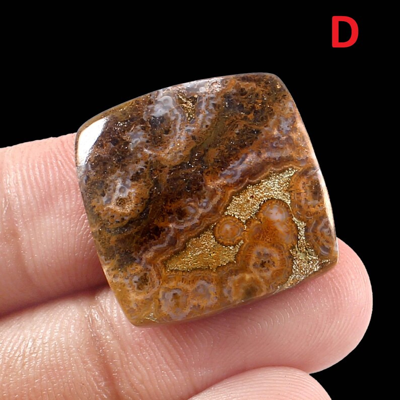 K&ouml;nnte beinhalten: Ein quadratischer, brauner und goldener Achat-Cabochon mit einem wirbelnden Muster. Der Stein hat eine wei&szlig;e, braune und goldene Farbgebung.