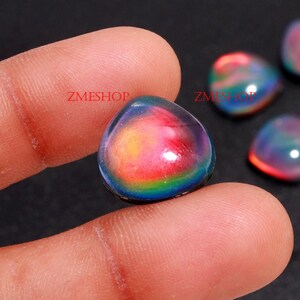 May include: A close-up of a iridescent gemstone cabochon with a rainbow of colors, including red, orange, yellow, green, blue, and purple. The gemstone is smooth and polished, and it is held in a person's hand.