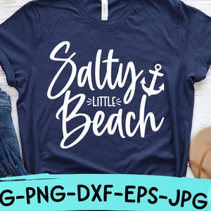 以下が含まれることがあります： 白い文字で「Salty Little Beach」と書いてあり、小さな錨のグラフィックが描かれたネイビーブルーのTシャツです。
