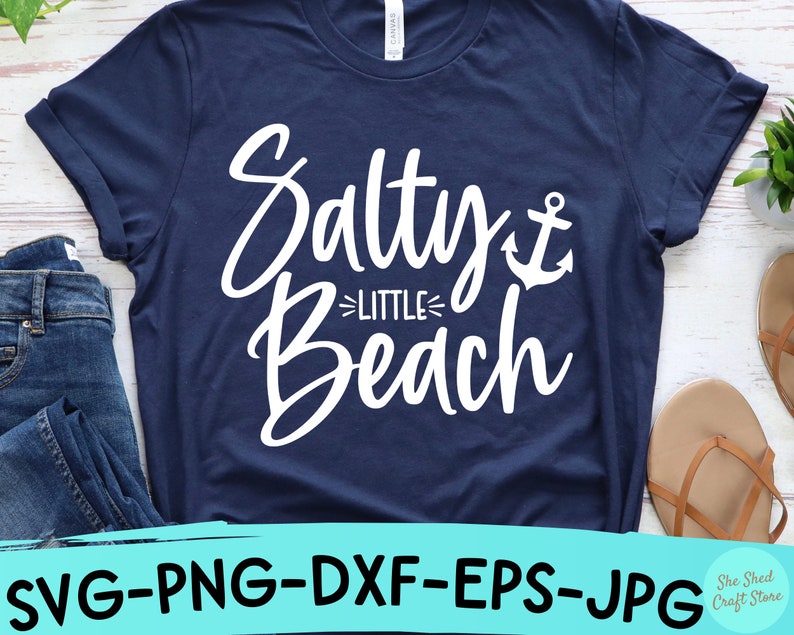 以下が含まれることがあります： 白い文字で「Salty Little Beach」と書いてあり、小さな錨のグラフィックが描かれたネイビーブルーのTシャツです。