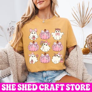 K&ouml;nnte beinhalten: Ein senfgelbes T-Shirt mit einer Rastergrafik aus neun Illustrationen. Das Design zeigt wei&szlig;e Geister mit rosa Schleifen und Blumen sowie rosa K&uuml;rbisse, die mit Schleifen verziert sind. Die Geister und K&uuml;rbisse sind in einem drei-mal-drei-Muster angeordnet.