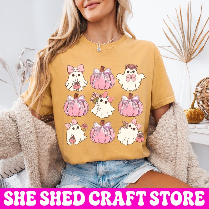 K&ouml;nnte beinhalten: Ein senfgelbes T-Shirt mit einer Rastergrafik aus neun Illustrationen. Das Design zeigt wei&szlig;e Geister mit rosa Schleifen und Blumen sowie rosa K&uuml;rbisse, die mit Schleifen verziert sind. Die Geister und K&uuml;rbisse sind in einem drei-mal-drei-Muster angeordnet.