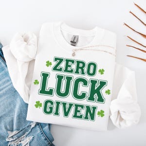 May include: White sweatshirt with the text "ZERO LUCK GIVEN" in green, along with green shamrock graphics. A silver necklace with a pendant is draped on the shirt. The sweatshirt is folded on top of blue denim.