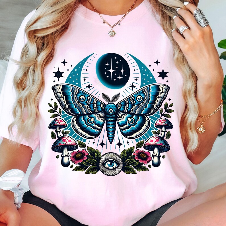 May include: A pink t-shirt with a graphic of a blue and gray moth with a crescent moon and stars in the background. The moth is surrounded by flowers and mushrooms. There is an eye in the center of the design.