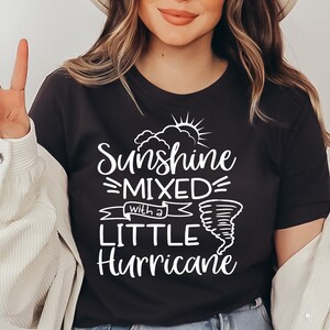 May include: Dark gray t-shirt with the text "Sunshine Mixed with a Little Hurricane" in white. The design includes a sun, clouds, and a tornado graphic. The shirt is a casual wear item.