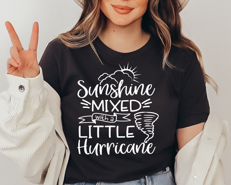 May include: Dark gray t-shirt with the text "Sunshine Mixed with a Little Hurricane" in white. The design includes a sun, clouds, and a tornado graphic. The shirt is a casual wear item.
