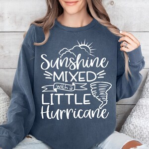 May include: A navy blue sweatshirt with the text "Sunshine Mixed with a Little Hurricane" in white. The design includes a sun, clouds, and a tornado graphic. The sweatshirt is a casual style.