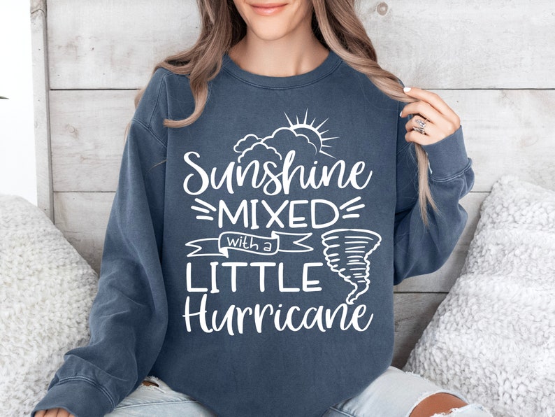 May include: A navy blue sweatshirt with the text "Sunshine Mixed with a Little Hurricane" in white. The design includes a sun, clouds, and a tornado graphic. The sweatshirt is a casual style.