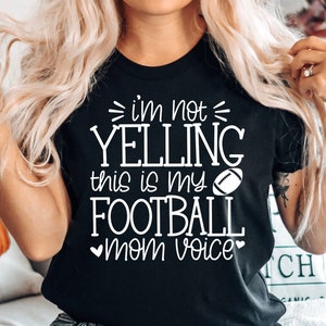 May include: Black t-shirt with white text that reads "i'm not yelling this is my football mom voice" with a white football graphic.