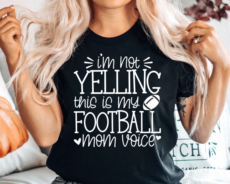 May include: Black t-shirt with white text that reads "i'm not yelling this is my football mom voice" with a white football graphic.