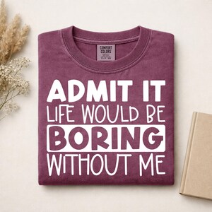 May include: A maroon crewneck t-shirt featuring the white text "ADMIT IT LIFE WOULD BE BORING WITHOUT ME" printed on the front. The shirt has a Comfort Colors brand label inside the collar and a ribbed neckline.