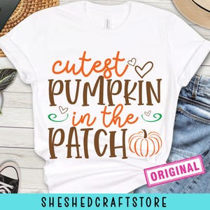 May include: White t-shirt with the text "cutest PUMPKIN in the PATCH" in orange and brown. The design includes a pumpkin graphic and hearts. A pink "ORIGINAL" sticker is visible. The bottom of the shirt has the text "SHESHEDCRAFTSTORE".
