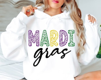 Puede incluir: Sudadera blanca con un diseño de texto negro "Mardi Gras". Las letras de "Mardi" están delineadas con estampado de leopardo verde, amarillo y morado.