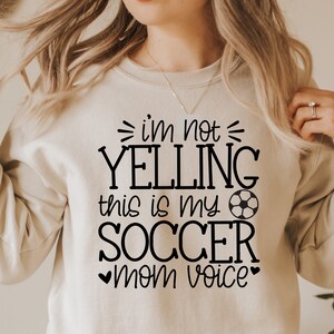 May include: A white sweatshirt with black text that reads "I'm not yelling this is my soccer mom voice" with a soccer ball graphic.