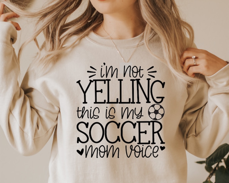 May include: A white sweatshirt with black text that reads "I'm not yelling this is my soccer mom voice" with a soccer ball graphic.