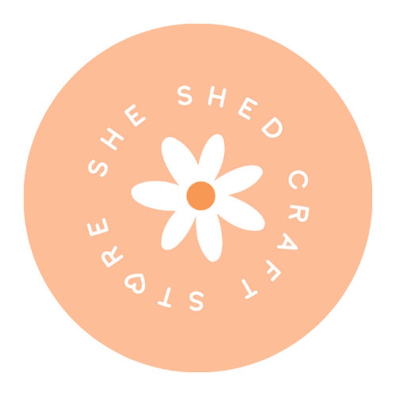 May include: A peach-colored circle with the text "She Shed Crafts" in white, surrounding a white daisy with a yellow center.