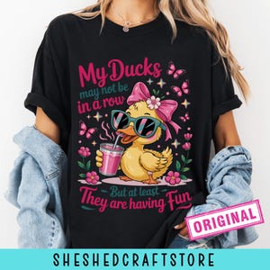 K&ouml;nnte beinhalten: Ein schwarzes T-Shirt mit einer Grafik einer gelben Ente, die eine t&uuml;rkisfarbene Sonnenbrille und eine rosa Schleife tr&auml;gt und ein rosa Getr&auml;nk h&auml;lt. Der Text lautet: My Ducks may not be in a row But at least They are having Fun. Rosa Blumen und Schmetterlinge umgeben die Ente.