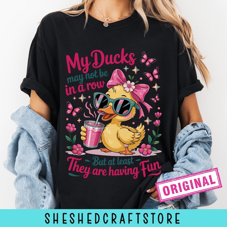 K&ouml;nnte beinhalten: Ein schwarzes T-Shirt mit einer Grafik einer gelben Ente, die eine t&uuml;rkisfarbene Sonnenbrille und eine rosa Schleife tr&auml;gt und ein rosa Getr&auml;nk h&auml;lt. Der Text lautet: My Ducks may not be in a row But at least They are having Fun. Rosa Blumen und Schmetterlinge umgeben die Ente.