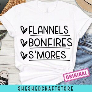 Op de afbeelding: Wit T-shirt met de zwarte woorden "FLANNELS BONFIRES S'MORES". Het shirt heeft een roze "ORIGINAL" tag en de winkelnaam "SHESHEDCRAFTSTORE" onderaan. Het shirt staat op een denim achtergrond.