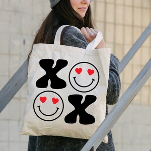 Pu&ograve; includere: Una borsa tote bianca con grafica nera e rossa. La grafica &egrave; composta da due facce sorridenti con cuori per gli occhi e le lettere "XO" in grassetto.