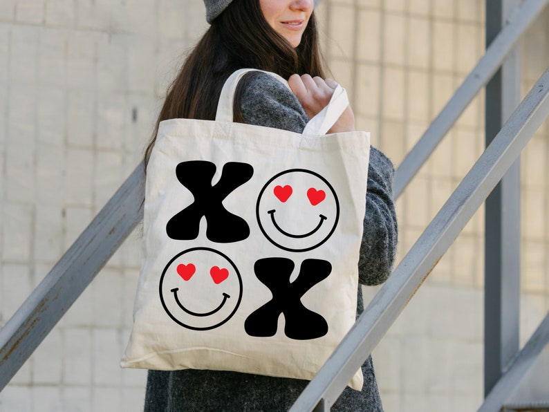Pu&ograve; includere: Una borsa tote bianca con grafica nera e rossa. La grafica &egrave; composta da due facce sorridenti con cuori per gli occhi e le lettere "XO" in grassetto.