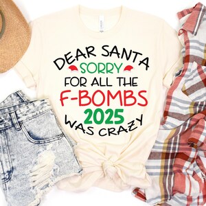 May include: Cream-colored t-shirt with the text "DEAR SANTA, SORRY FOR ALL THE F-BOMBS 2025 WAS CRAZY" in black, red, and green. The shirt is styled with denim shorts, a straw hat, and a red and white plaid shirt.