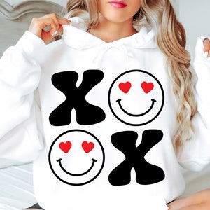 Pu&ograve; includere: Felpa con cappuccio bianca con un design grafico nero e rosso con le lettere "XO" e due facce sorridenti con cuori per occhi.